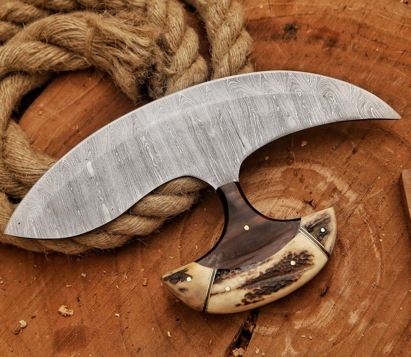 A curved Damascus steel knife with a patterned steel blade, dark brown handle, and cream bone inlay, displayed on a rustic wooden surface next to a coil of rope.