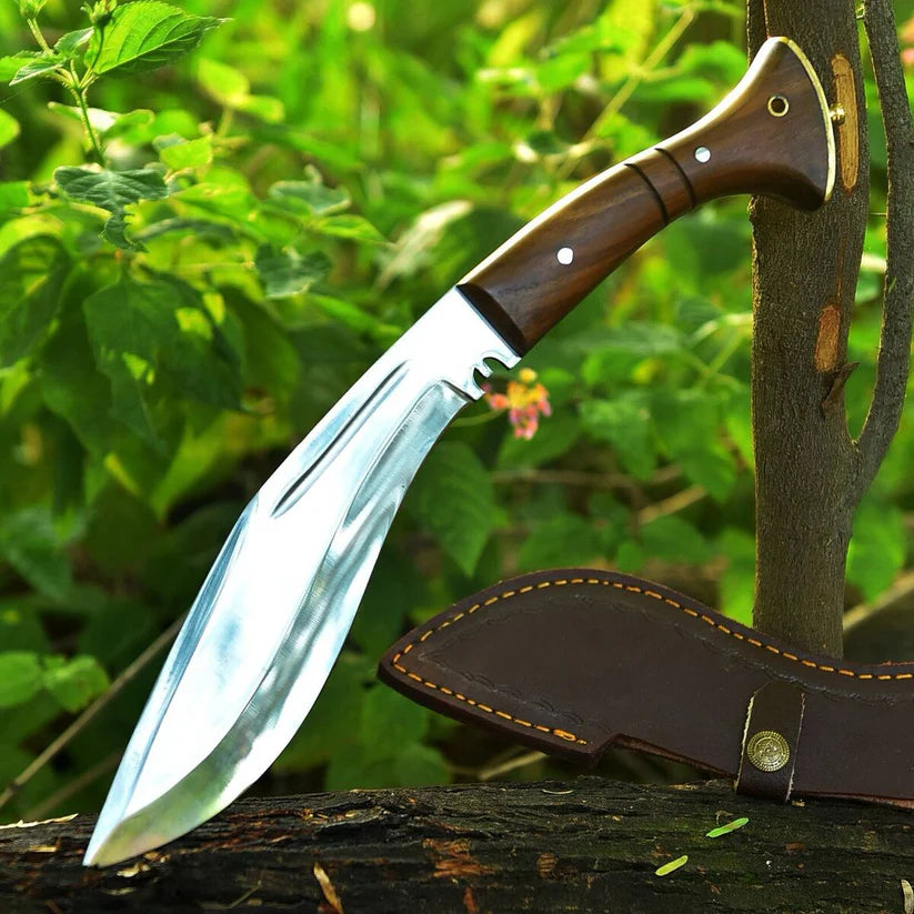 A large machete-style knife with a polished silver blade and a brown wooden handle, shown leaning against a tree trunk outdoors with a brown leather sheath beside it.