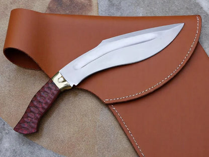 A fixed-blade knife with a polished silver blade, gold bolster, and reddish-brown textured handle, resting on a brown leather sheath over a tan and gray stone surface.
