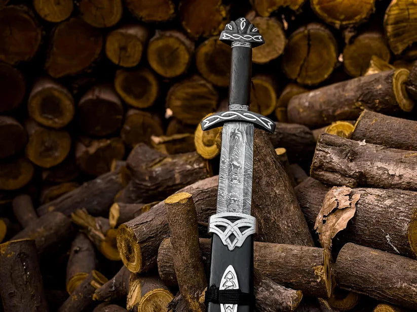 A decorative sword with a textured silver blade, black handle, and ornate silver guard and pommel, standing upright in a pile of cut logs.