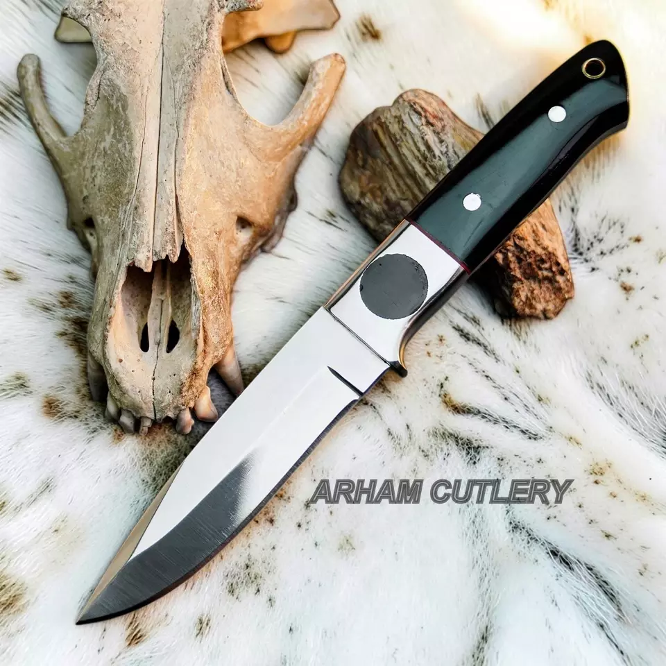 A fixed-blade knife with a polished silver blade and a black handle with silver rivets, laid diagonally on a white fur surface next to a deer skull and a rock. The text 'ARHAM CUTLERY' is overlaid on the image.