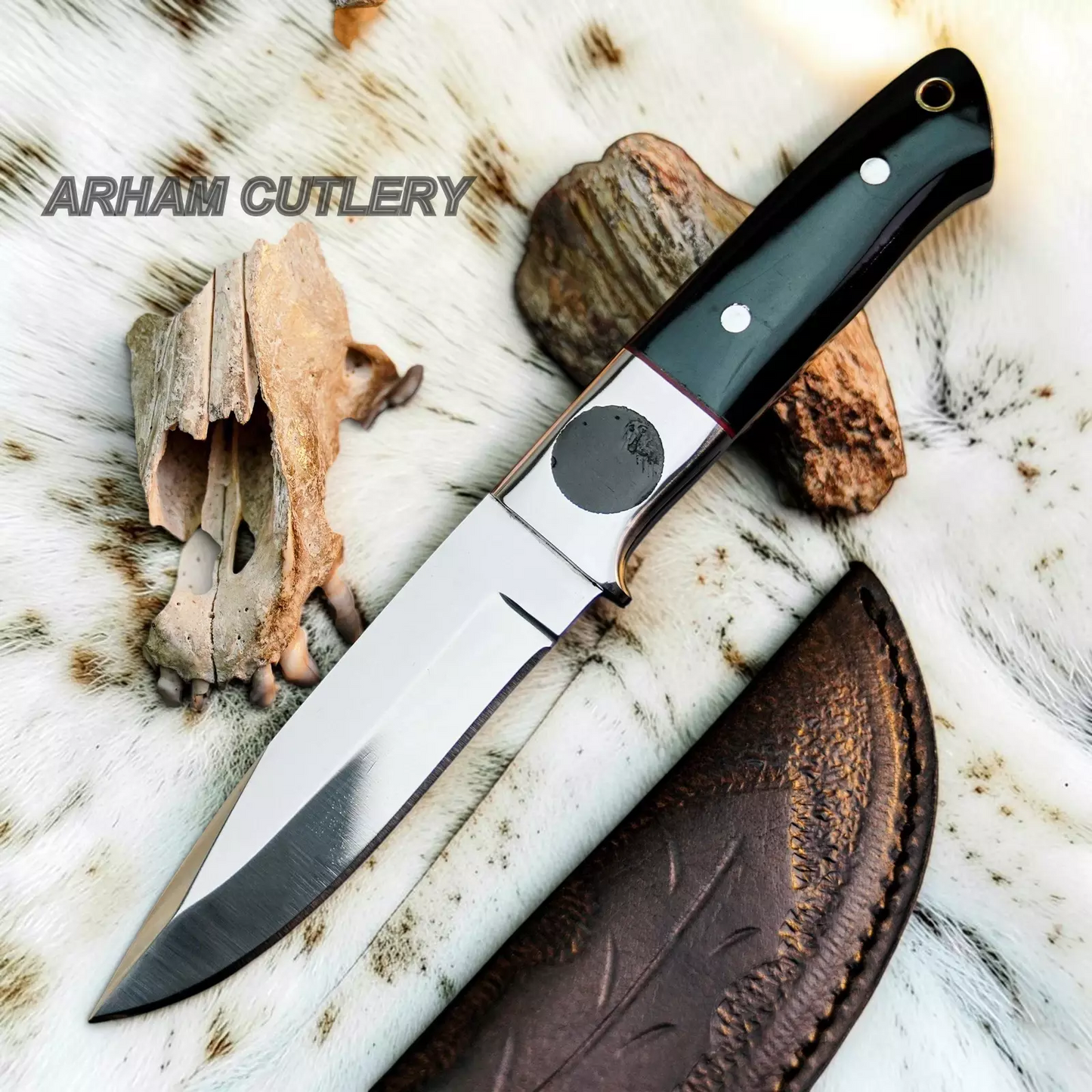 A fixed-blade knife with a polished steel blade, black handle scales, and silver rivets, displayed diagonally on a white fur background with animal bones, rocks, and a brown leather sheath. The text 'ARHAM CUTLERY' is visible in the upper left.