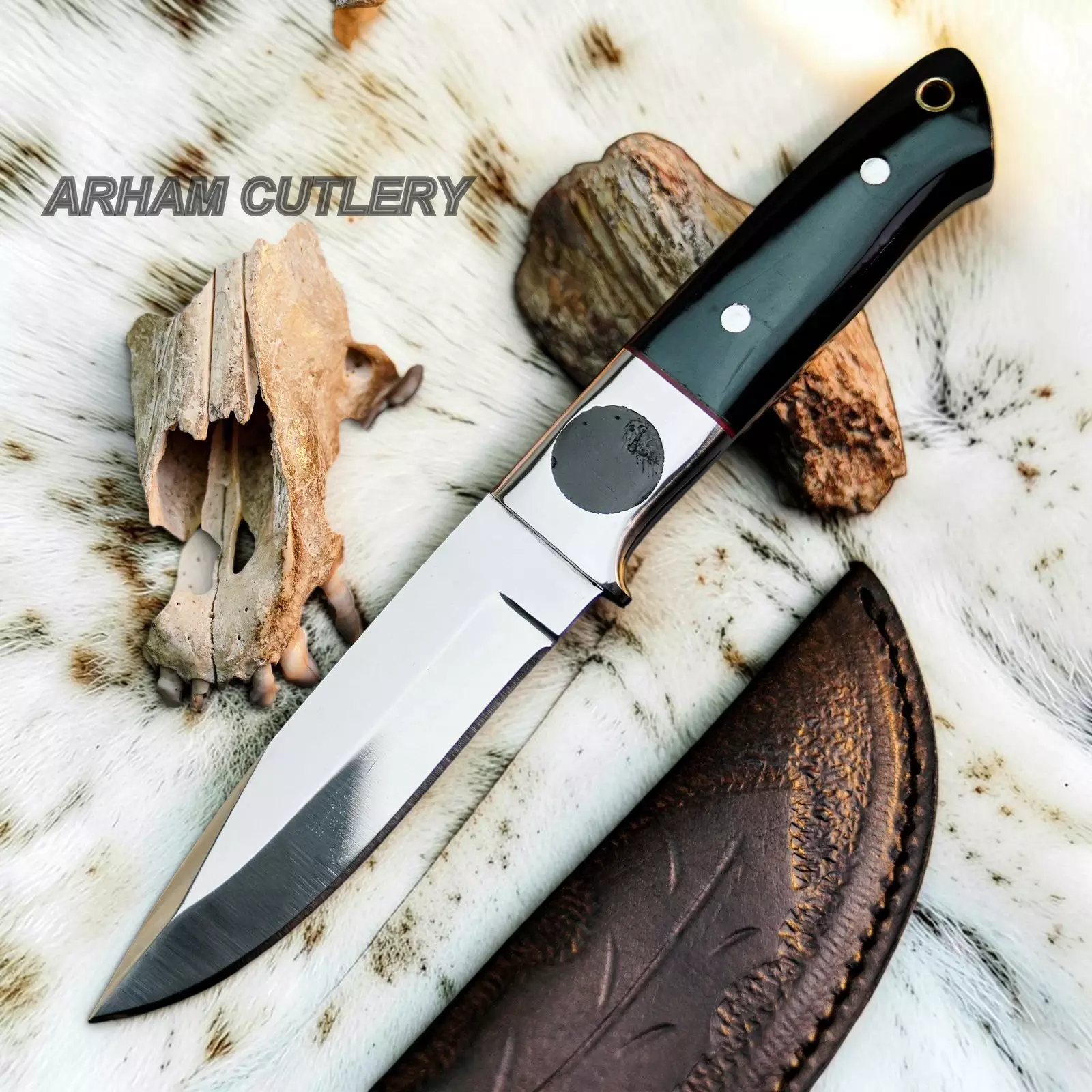 A fixed-blade knife with a polished steel blade, black handle scales, and silver rivets, displayed diagonally on a white fur background with animal bones, rocks, and a brown leather sheath. The text 'ARHAM CUTLERY' is visible in the upper left.