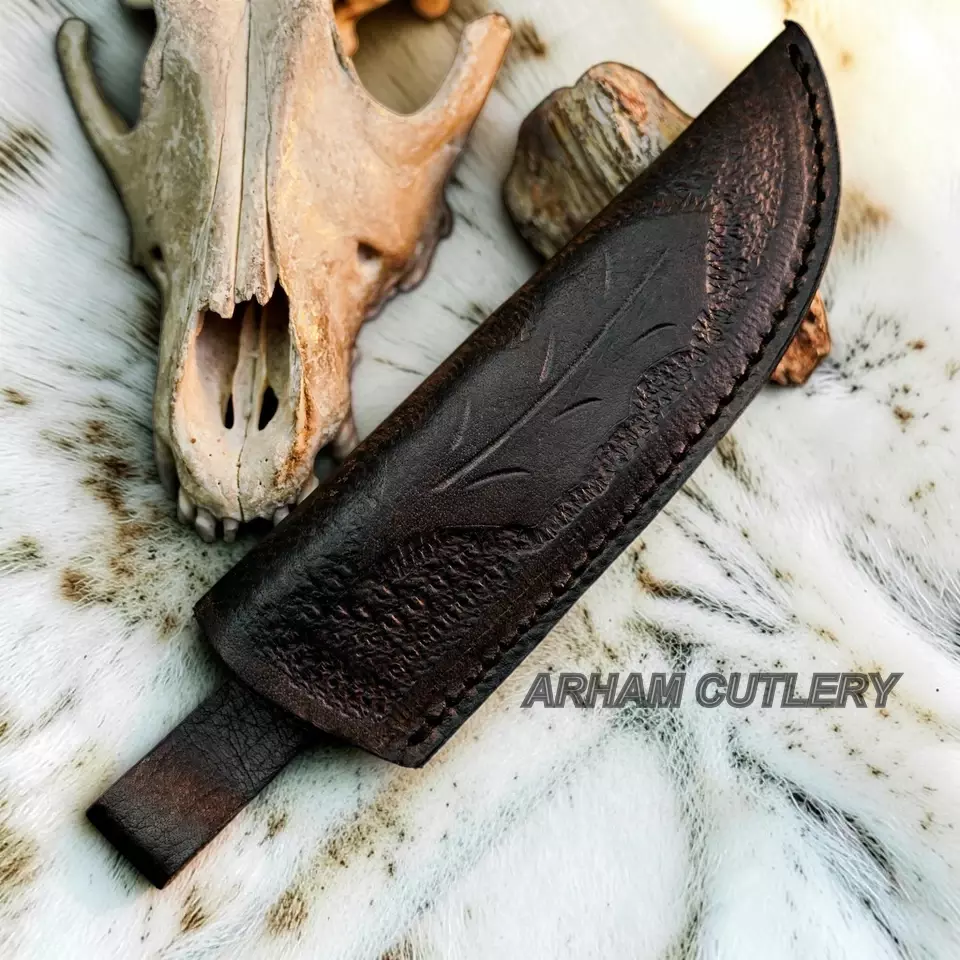 A dark brown leather knife sheath with embossed leaf detailing, shown on a white fur surface with a deer skull and wood prop. The text 'ARHAM CUTLERY' is overlaid on the image.