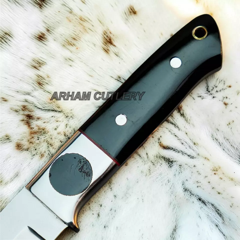 A close-up of a knife handle with a glossy black grip, silver bolster, and brass eyelet, resting on a white and brown spotted fur surface. A translucent 'ARHAM CUTLERY' watermark is visible across the center.