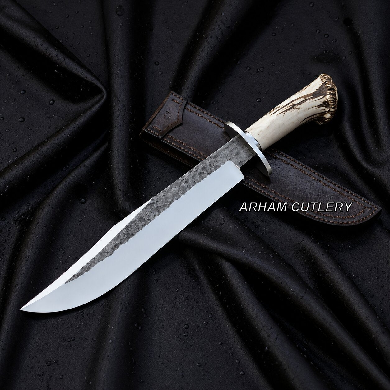 A fixed-blade knife with a hammered steel blade, silver bolster, and antler-style handle, resting diagonally on a dark brown leather sheath over a draped black satin fabric background. The text 'ARHAM CUTLERY' is overlaid on the right side.