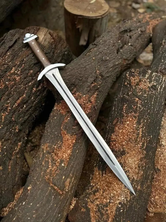 A medieval-style short sword with a silver blade, white crossguard, and brown handle, resting diagonally on dark weathered logs outdoors.