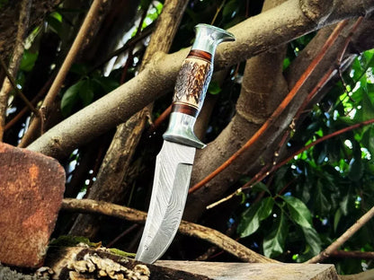 A custom fixed-blade knife with a patterned steel blade, a carved wooden handle with bone inlay, and a polished silver guard, shown standing upright on a log surrounded by tree branches and leaves.