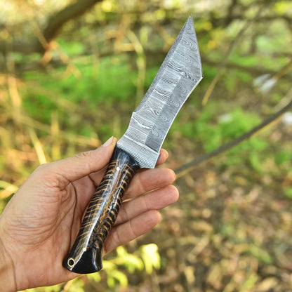 A hand holding a fixed-blade knife with a patterned Damascus-style steel blade and a dark brown and black striped handle, set against a blurred outdoor forest background.