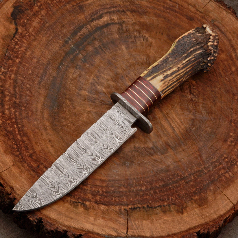 A fixed-blade knife with a patterned Damascus steel blade, a brown leather-wrapped guard, and a natural antler handle, displayed diagonally on a rustic wood slice background.