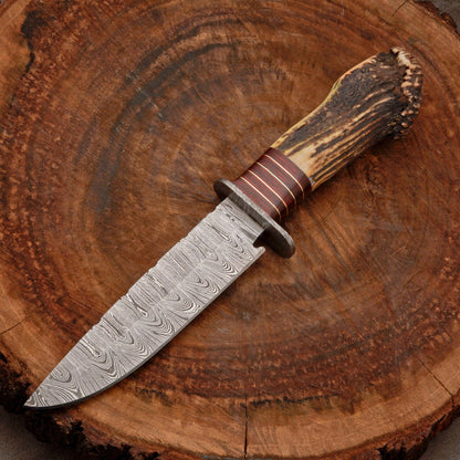A fixed-blade knife with a patterned Damascus steel blade, a brown leather-wrapped guard, and a natural antler handle, displayed diagonally on a rustic wood slice background.