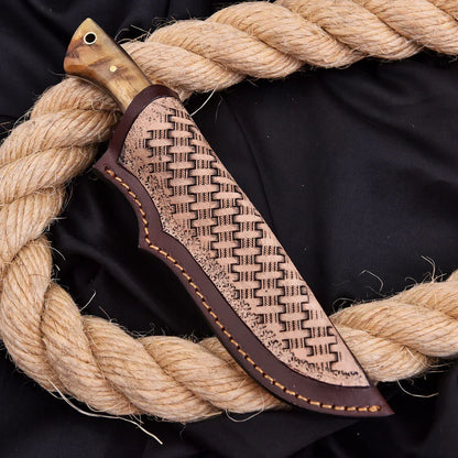 A close-up of a knife in a decorative brown leather sheath, arranged on a black fabric background with a large coiled rope around it. The knife handle appears wood-toned, and the sheath has a woven or embossed pattern with visible contrast stitching.