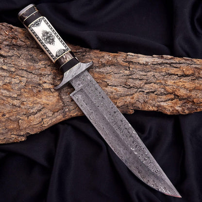 A custom handmade knife with a Damascus steel blade and an ornate handle, resting on a piece of tree bark over a black fabric background.