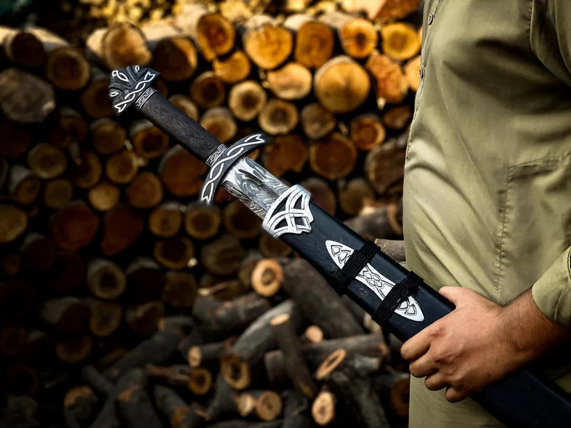 A person in a khaki shirt holds a black and silver decorative sword or dagger in front of a stacked background of cut logs.