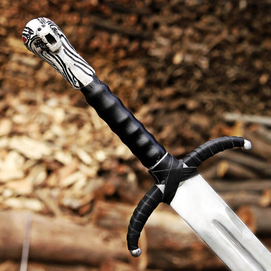 A close-up of a decorative sword with a black wrapped handle, a carved silver pommel, and a silver blade, set against a blurred background of wood and leaves.