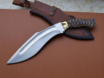 A fixed-blade knife with a polished silver blade, gold-toned bolster, and a carved dark wood handle, displayed next to its brown leather sheath on a brown leather surface.