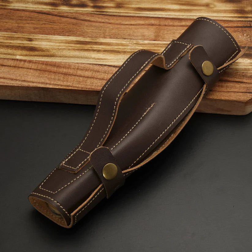 A dark brown leather knife sheath with tan stitching and brass snap closures, resting diagonally on a dark gray surface with a wooden board in the background.