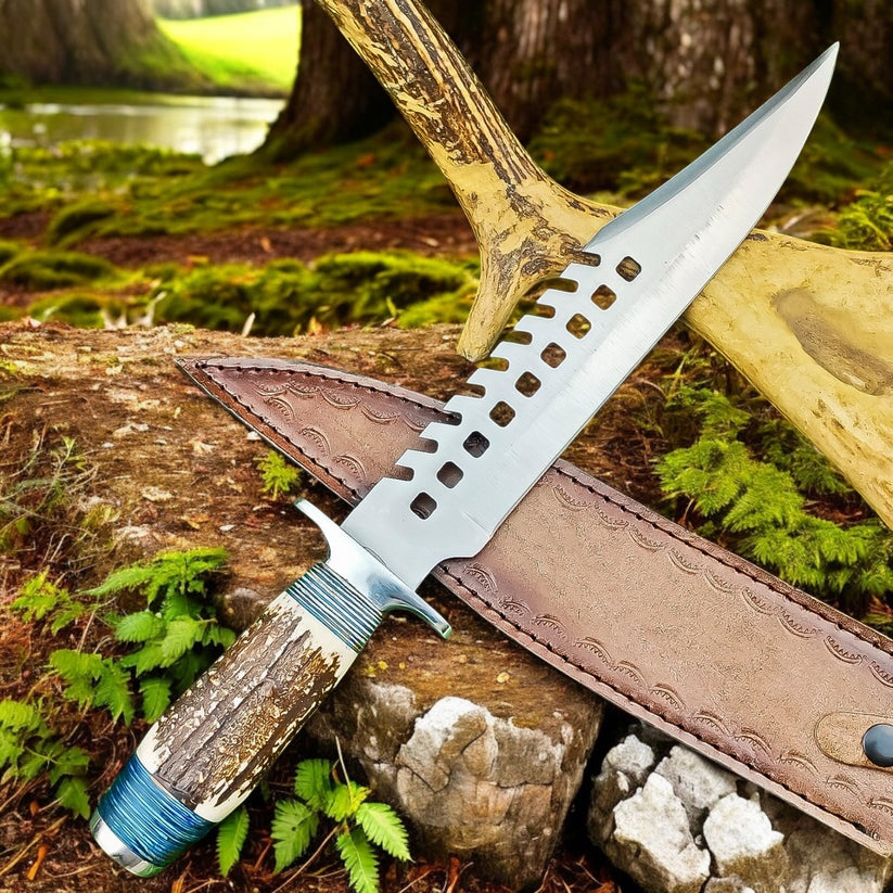 A fixed-blade knife with a textured antler handle and a silver blade featuring a decorative cutout pattern, displayed diagonally on a brown leather sheath outdoors on a mossy log with trees and ferns in the background.