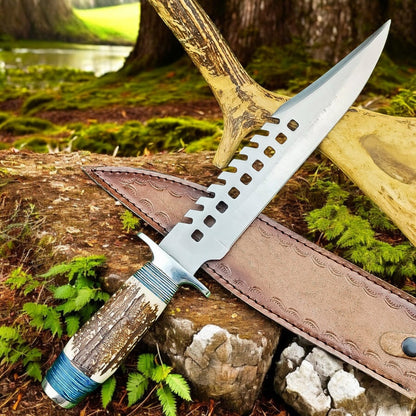A fixed-blade knife with a textured antler handle and a silver blade featuring a decorative cutout pattern, displayed diagonally on a brown leather sheath outdoors on a mossy log with trees and ferns in the background.