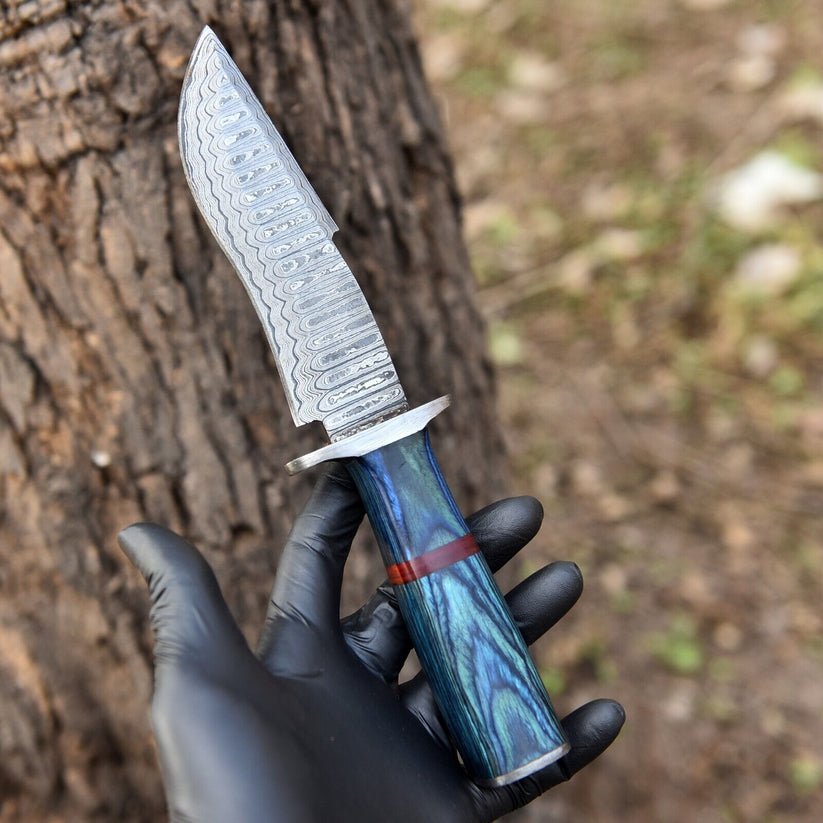 A hand wearing a black glove holds a fixed-blade knife with a patterned steel blade and a blue marbled handle with a red accent band, set against a blurred tree trunk and outdoor background.