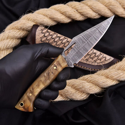 A hand wearing a black glove holds a fixed-blade knife with a patterned steel blade and a carved wooden handle, positioned in front of a woven sheath and thick natural rope on a black fabric background.
