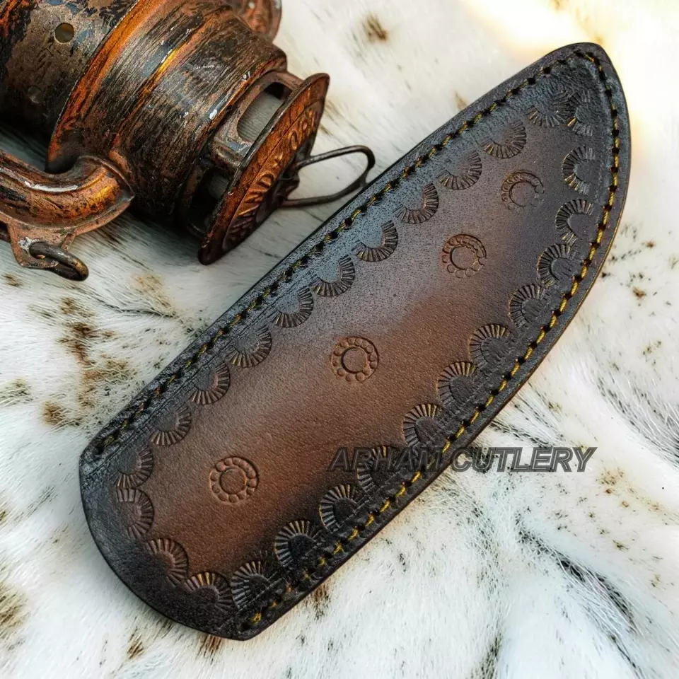 A dark brown leather knife sheath with hand-tooled circular patterns and yellow stitching, displayed on a white fur surface next to an antique wooden object. A watermark for 'ABAM CUTLERY' is visible.