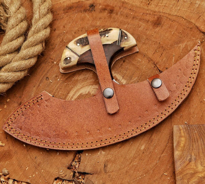 A curved leather sheath with a snap strap holding a carved wooden and bone-handled knife, displayed on a rustic wood surface with rope and wood chips nearby.