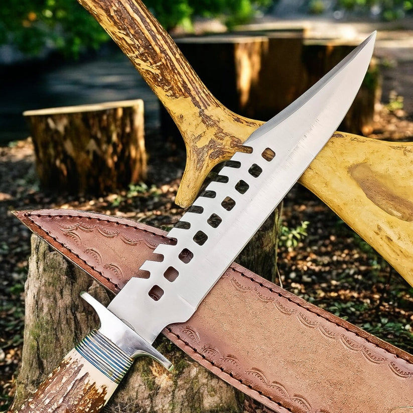 A fixed-blade knife with a silver blade featuring a distinctive square-hole cutout pattern along the spine, resting on a brown leather sheath. The knife is set against a rustic outdoor background with tree stumps and a large antlered axe handle.