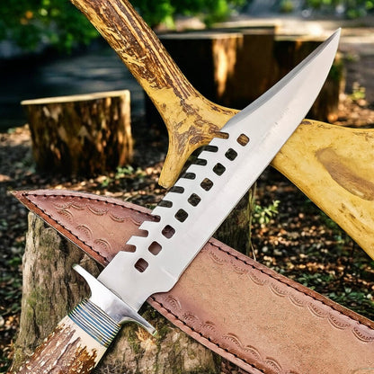 A fixed-blade knife with a silver blade featuring a distinctive square-hole cutout pattern along the spine, resting on a brown leather sheath. The knife is set against a rustic outdoor background with tree stumps and a large antlered axe handle.