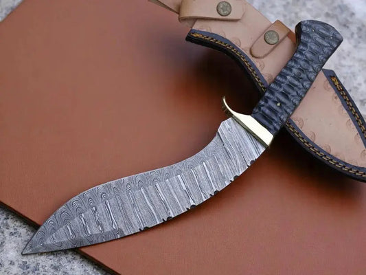 A curved fixed-blade knife with a patterned Damascus steel blade, a dark textured handle, and a brass guard, resting on a brown leather sheath over a brown leather surface.