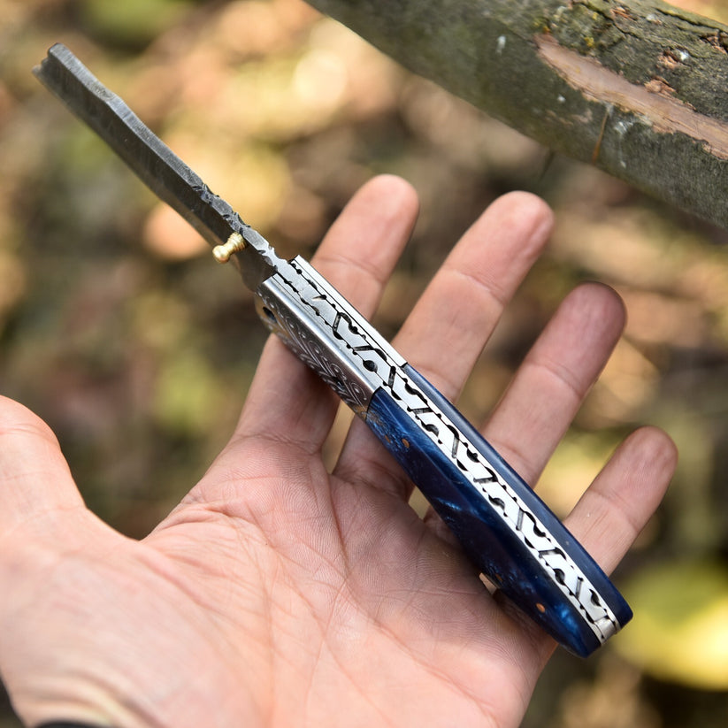 A folding pocket knife with a dark blue marbled handle and silver engraved metal accents, held in a person's hand against a blurred outdoor background.