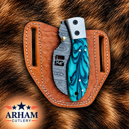A folding knife with a turquoise and blue marbled handle and a gray patterned blade, displayed in a brown leather sheath on a brown fur background. The ARHAM CUTLERY logo is visible in the bottom-left corner.