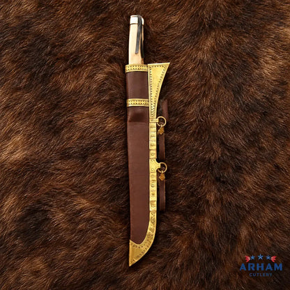 A decorative knife with a polished silver-toned blade and ornate gold-toned guard, shown partially sheathed in a brown leather sheath with matching gold trim, laid on a brown fur background. The Arham Cutlery logo is visible in the bottom right corner.