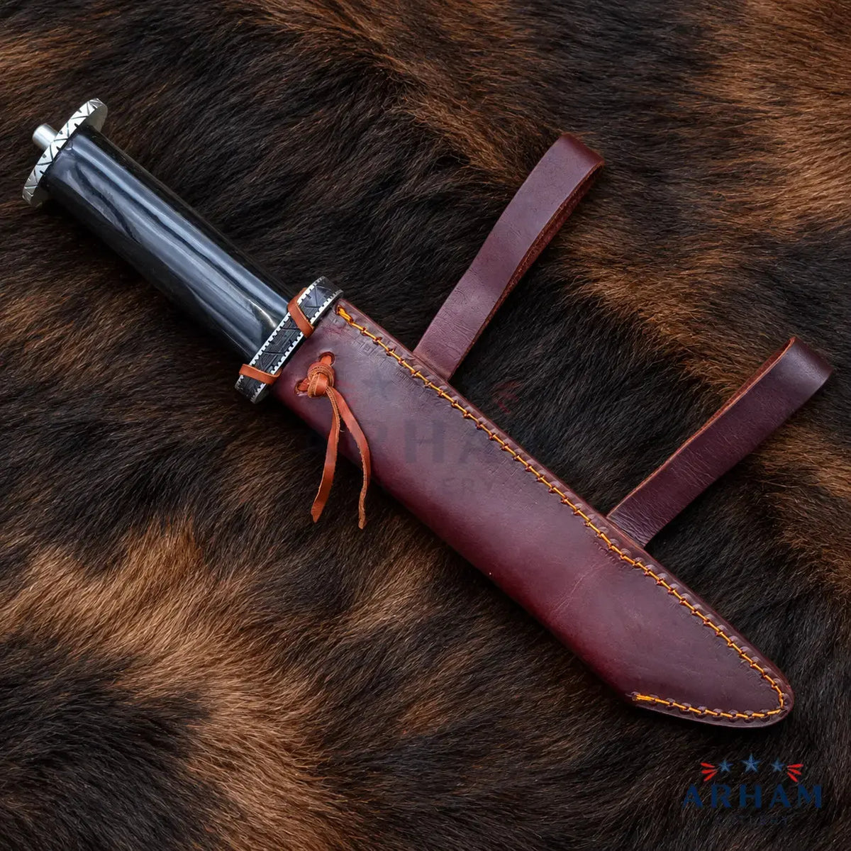 A decorative dagger with a black handle and silver guard, resting diagonally on a brown and black animal fur surface next to its reddish-brown leather sheath with orange stitching.