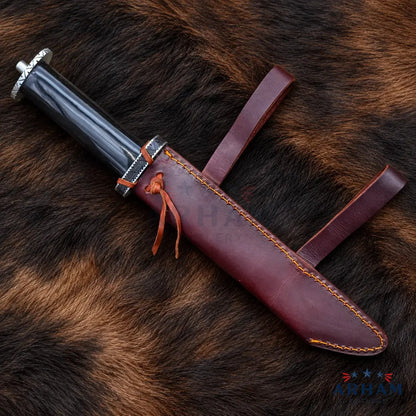 A decorative dagger with a black handle and silver guard, resting diagonally on a brown and black animal fur surface next to its reddish-brown leather sheath with orange stitching.