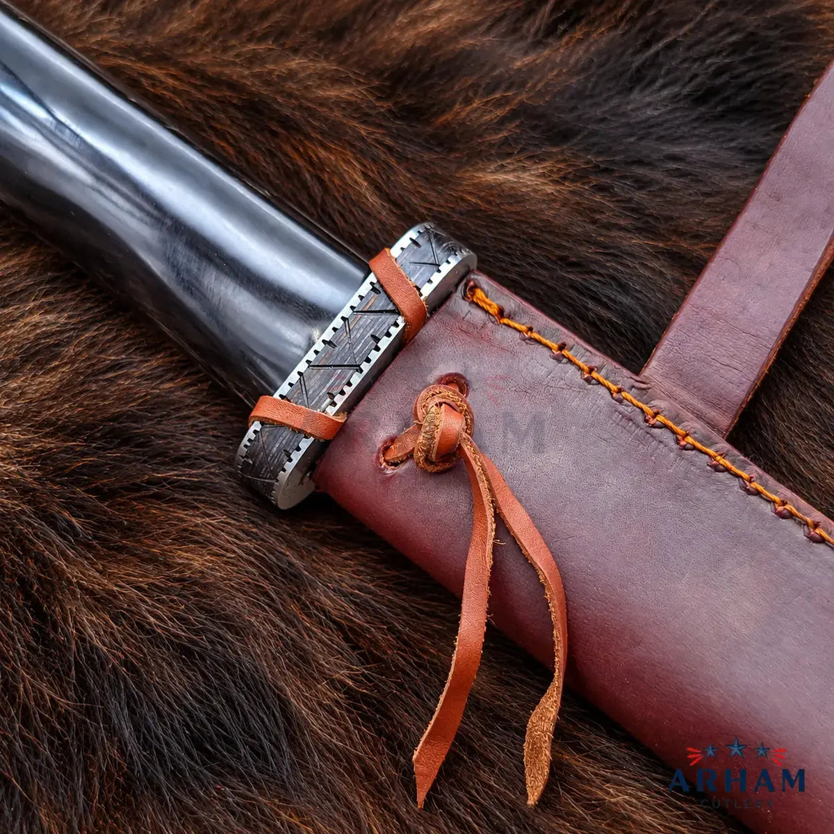 A close-up of a dark metal knife or dagger handle with a silver engraved guard, wrapped with brown leather cords, resting on a brown fur surface next to a brown leather sheath with orange stitching and a leather tie. The Alham brand logo is visible in the bottom right corner.
