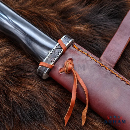 A close-up of a dark metal knife or dagger handle with a silver engraved guard, wrapped with brown leather cords, resting on a brown fur surface next to a brown leather sheath with orange stitching and a leather tie. The Alham brand logo is visible in the bottom right corner.
