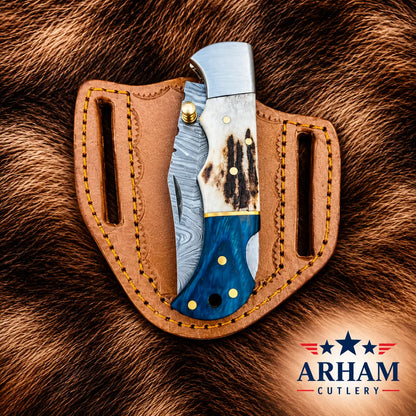 A folding pocket knife with a Damascus steel blade, a white and black bone handle, and a blue wood inlay, displayed in a brown leather sheath on a brown fur background. The ARHAM CUTLERY logo is visible in the bottom right corner.