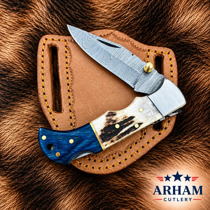 A folding pocket knife with a patterned steel blade, a bone handle, and a blue wood handle is displayed on a brown leather sheath over a brown fur background. The ARHAM CUTLERY logo is visible in the bottom right corner.