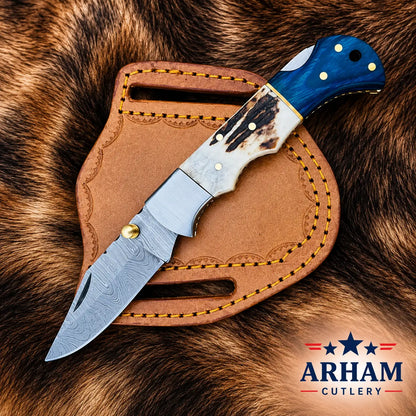 A folding pocket knife with a Damascus steel blade, a weathered ivory handle, and a blue contoured handle is displayed diagonally on a brown leather sheath, all set against a brown fur background. The Arham Cutlery logo is visible in the bottom right corner.