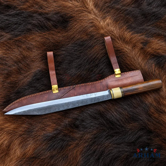 A fixed-blade knife with a patterned steel blade, dark wood handle, and brass guard, displayed diagonally on its brown leather sheath over a brown fur background. The brand logo 'ARTHAM' is visible in the bottom right corner.