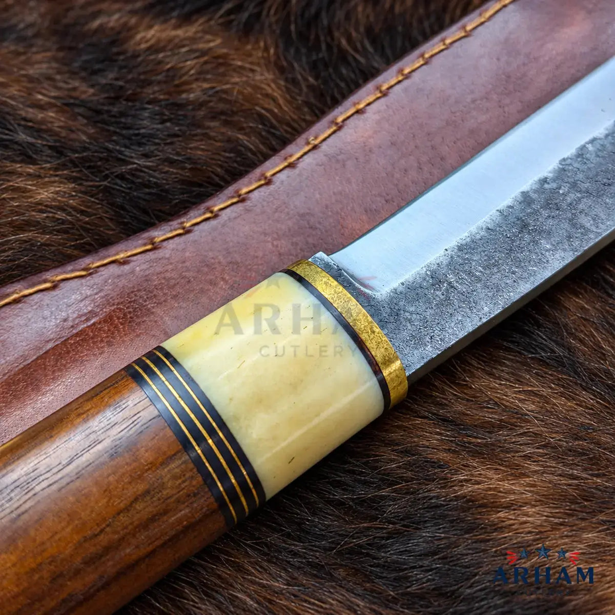 A close-up of a fixed-blade knife with a polished wood handle, a textured steel blade, and a cream-colored inlay ring with gold trim, resting on a brown leather sheath over a dark brown fur background. A semi-transparent 'ARH CUTLERY' watermark is visible across the handle, and a 'ARH' logo is in the bottom right corner.