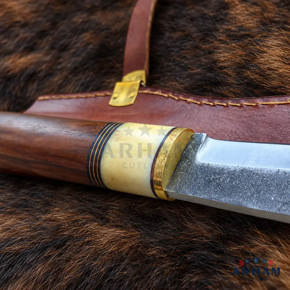 A close-up of a fixed-blade knife with a polished dark wood handle, a silver steel blade, and brass and ivory-colored guard details, resting on a brown leather sheath over a dark brown fur background.