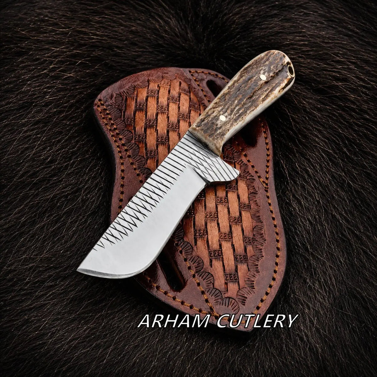 A fixed-blade knife with a silver patterned blade and a brown antler-style handle, resting diagonally on a brown woven leather sheath over a dark fur background. The text 'ARHAM CUTLERY' is printed at the bottom.