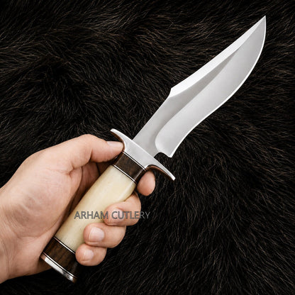 A hand holding a fixed-blade knife with a polished silver blade, a cream-colored bone handle, and dark brown wood accents, set against a black fur background. The brand name 'ARHAM CUTLERY' is watermarked across the center.