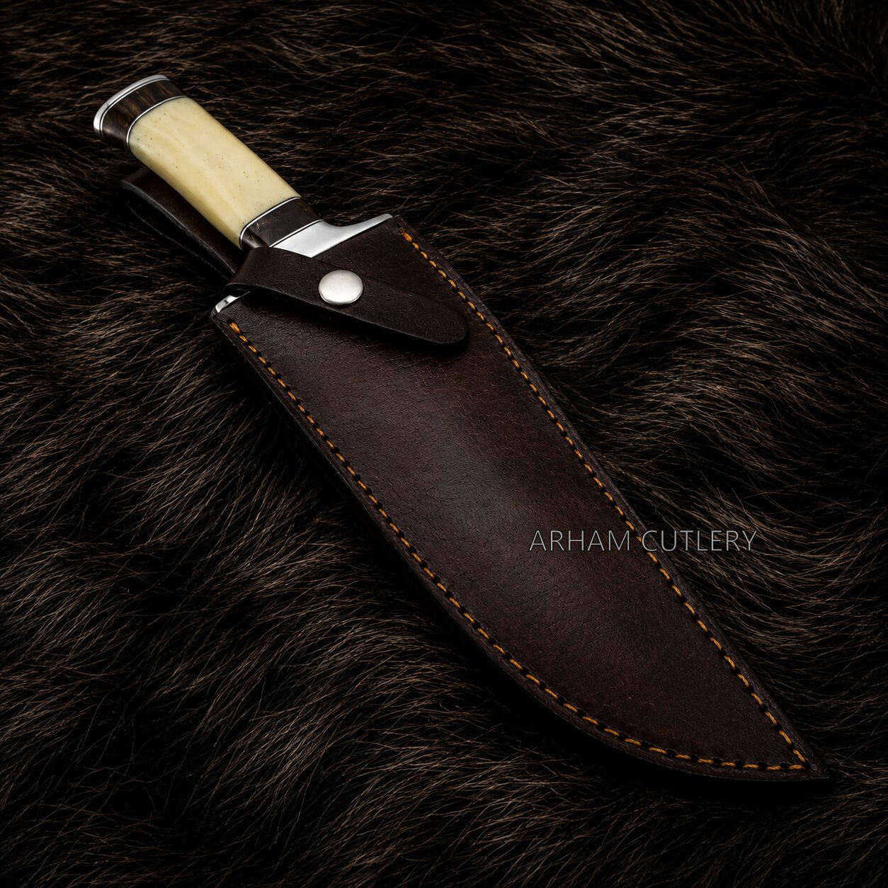 A fixed-blade knife with a cream-colored handle and silver bolster, shown partially sheathed in a dark brown leather sheath with contrast stitching, placed on a dark brown fur background. The brand name 'ARHAM CUTLERY' is watermarked on the sheath.