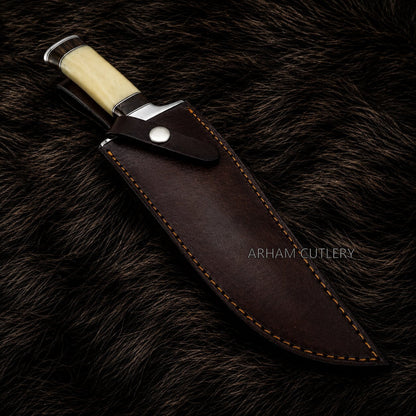 A fixed-blade knife with a cream-colored handle and silver bolster, shown partially sheathed in a dark brown leather sheath with contrast stitching, placed on a dark brown fur background. The brand name 'ARHAM CUTLERY' is watermarked on the sheath.