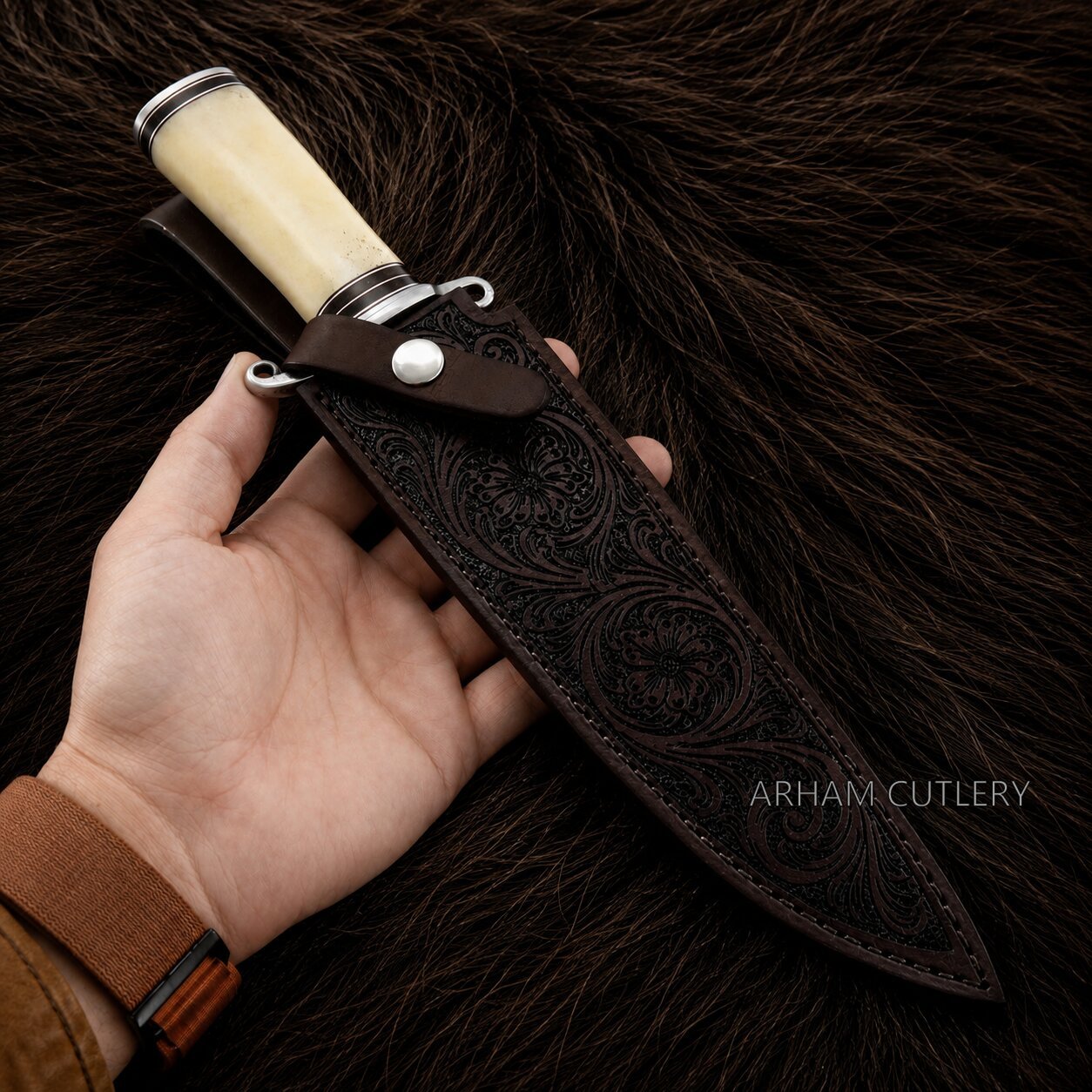 A hand holds a fixed-blade knife with a cream-colored handle and a dark brown leather sheath featuring embossed floral patterns, set against a dark brown fur background. The brand name 'ARHAM CUTLERY' is visible as a watermark.