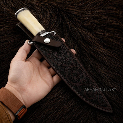 A hand holds a fixed-blade knife with a cream-colored handle and a dark brown leather sheath featuring embossed floral patterns, set against a dark brown fur background. The brand name 'ARHAM CUTLERY' is visible as a watermark.