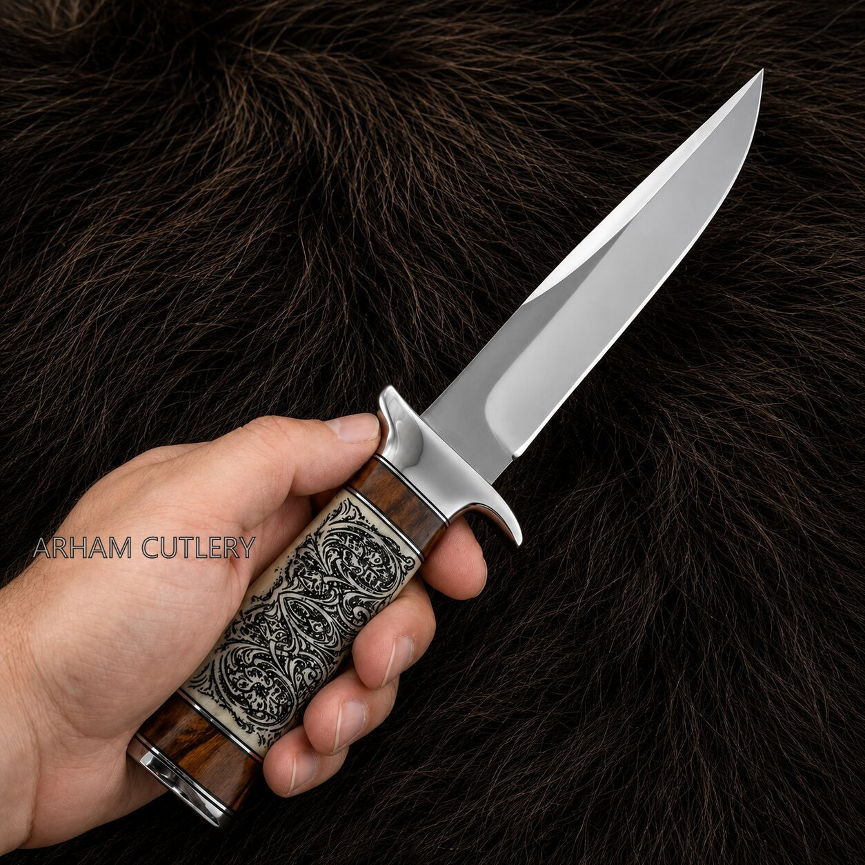 A hand holding a fixed-blade knife with a polished steel blade and an intricately carved brown and silver handle, set against a dark brown fur background. The watermark 'ARHAM CUTLERY' is visible on the left.